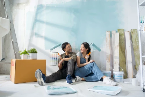 Couple sitting on the floor with paint rollers, enjoying painting a room together.