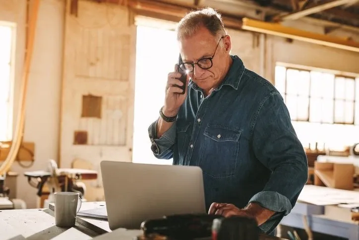 Man standing in a workshop looks down at a laptop while on the phone.