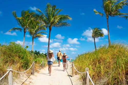 Group of people walking down a beach path on a sunny day with palm trees.